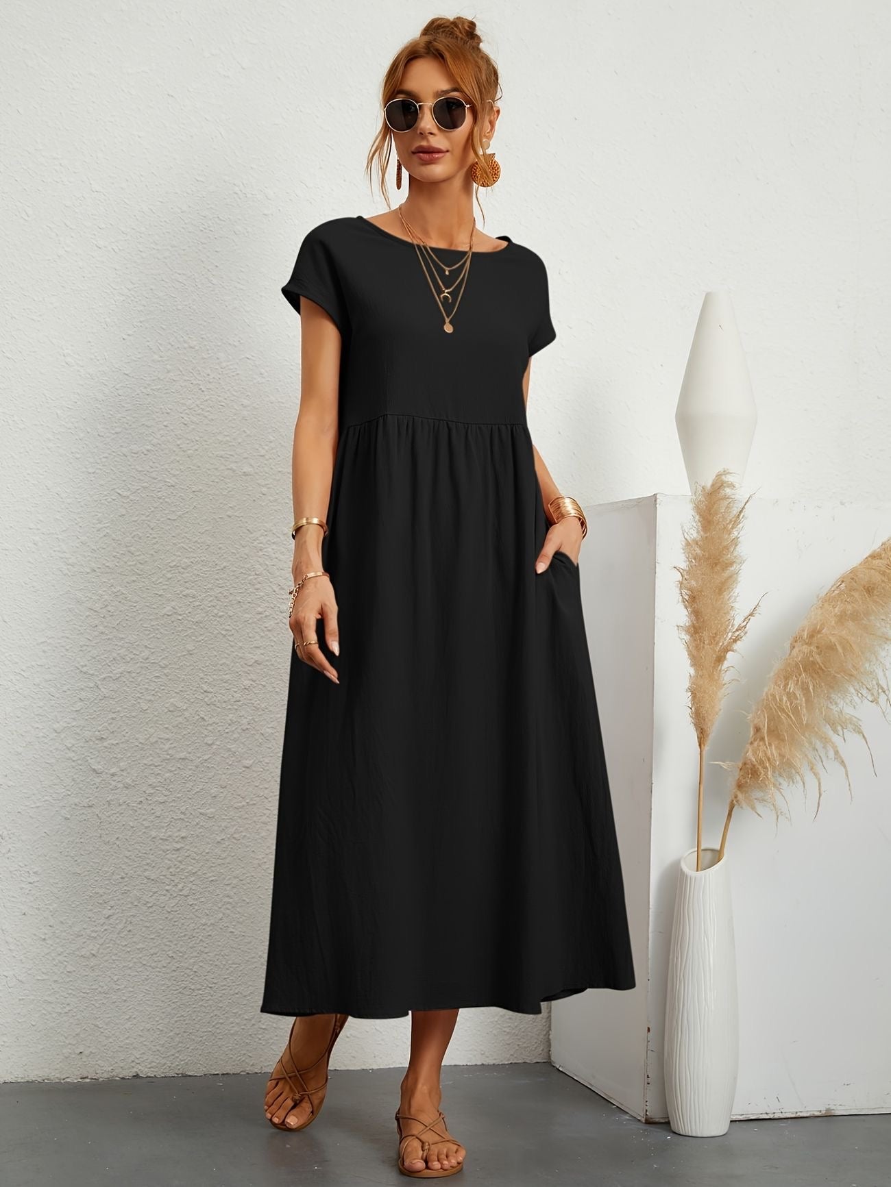 Donna con abito lungo nero casual, occhiali da sole, sandali marroni, accessori dorati, accanto a vaso bianco con pampas. Moda estiva elegante.