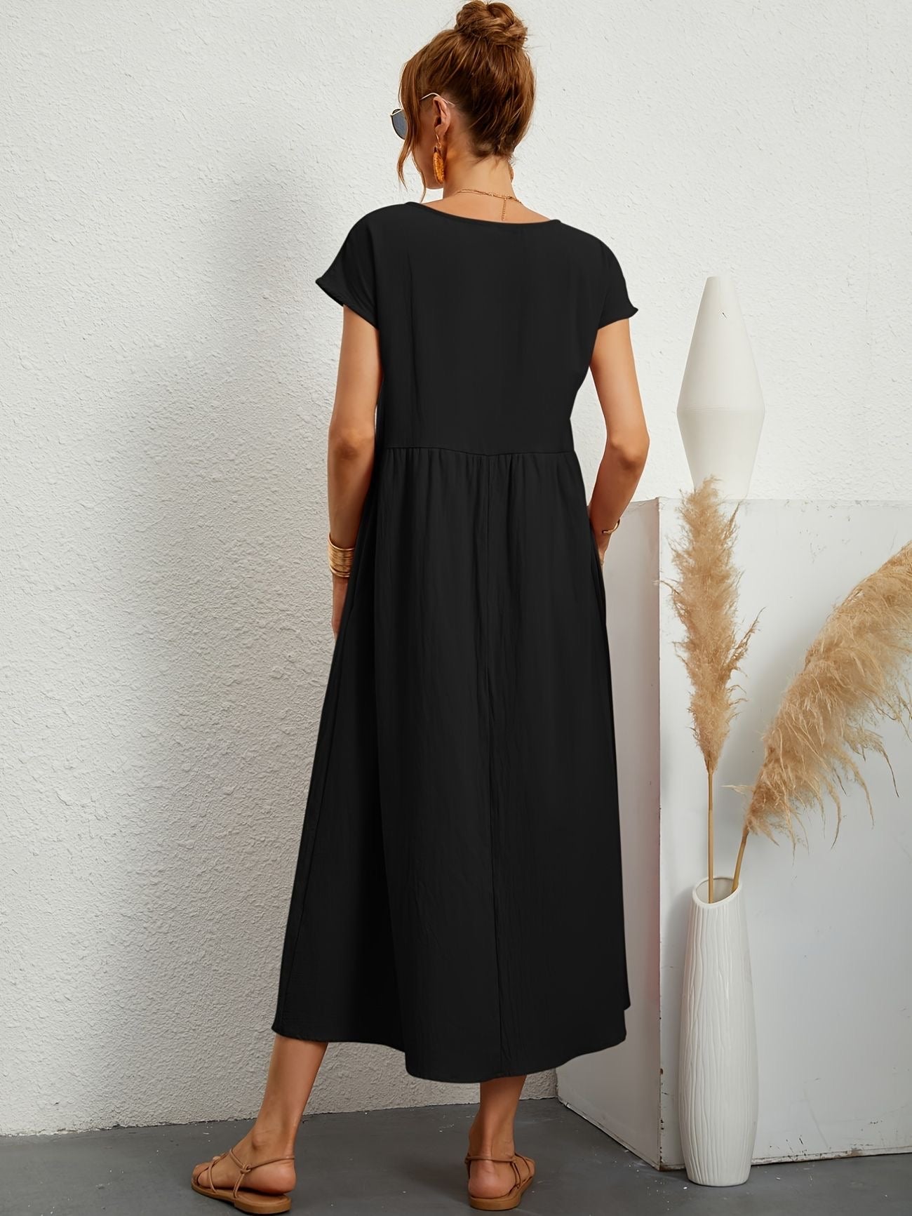 Donna con abito lungo nero, sandali beige, capelli raccolti, occhiali da sole, accanto a vaso bianco con pampas. Moda estiva elegante e casual.