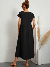 Donna con abito lungo nero, sandali beige, capelli raccolti, occhiali da sole, accanto a vaso bianco con pampas. Moda estiva elegante e casual.