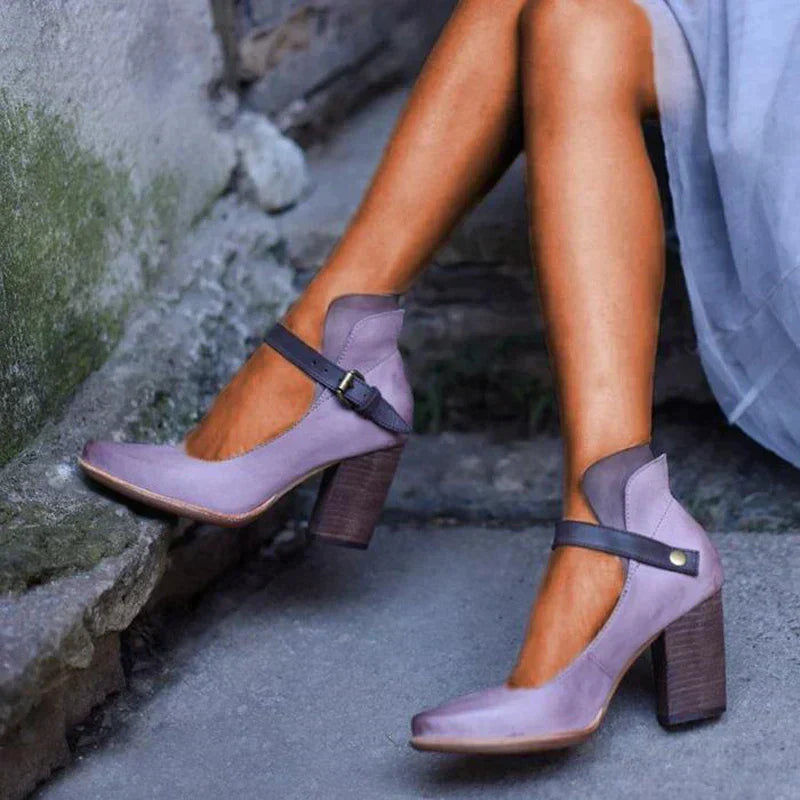 Scarpe col tacco alto viola con cinturino nero, indossate da una donna con abito grigio. Design elegante, perfette per moda autunnale.