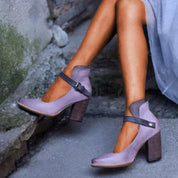 Scarpe col tacco alto viola con cinturino nero, indossate da una donna con abito grigio. Design elegante, perfette per moda autunnale.