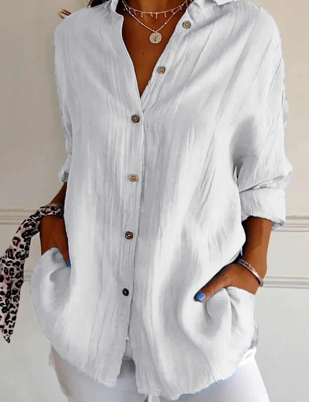 Camicia bianca da donna in lino, maniche lunghe, bottoni marroni, tasche frontali, stile casual elegante, abbinata a pantaloni bianchi.