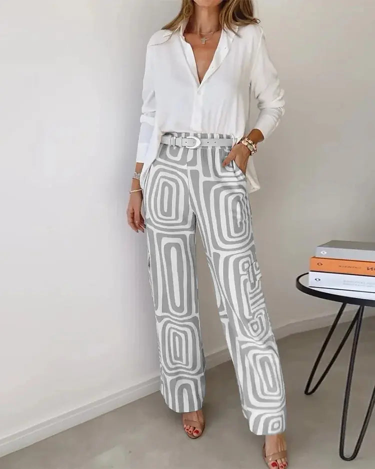 Donna con camicia bianca e pantaloni a palazzo grigi con motivo geometrico, in piedi accanto a un tavolo moderno con libri. Moda elegante e contemporanea.