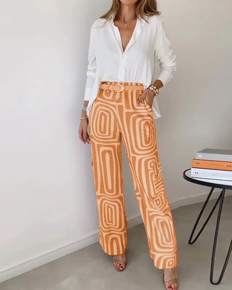 Donna in camicia bianca e pantaloni arancioni a motivi geometrici, in piedi accanto a un tavolo con libri. Moda casual elegante, abbigliamento femminile.