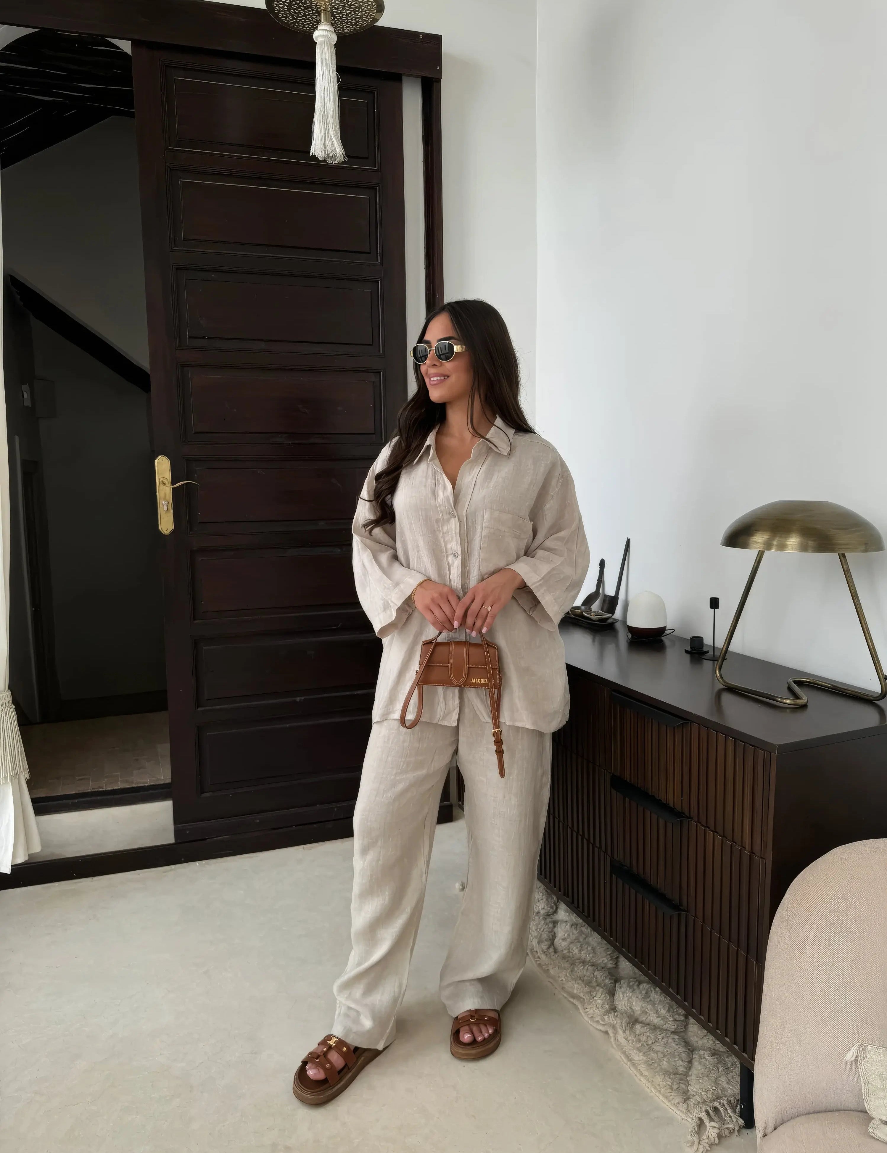Donna con occhiali da sole indossa completo beige in lino, borsa a tracolla marrone, sandali, in soggiorno moderno con mobili in legno scuro.