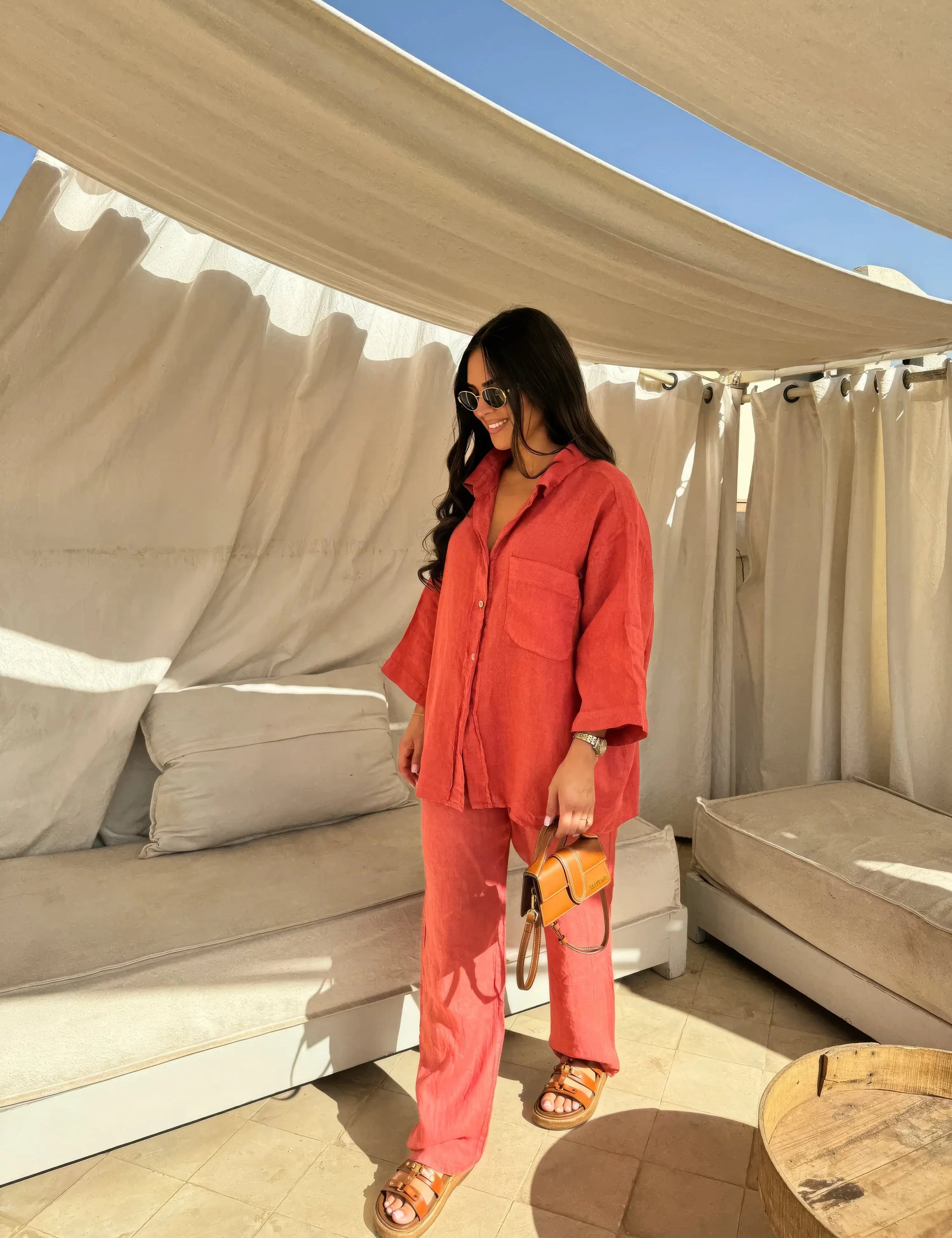Donna con camicia e pantaloni rossi, occhiali da sole, borsa a tracolla marrone, sandali, in terrazza con tende bianche. Moda estiva casual.