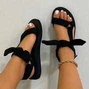 Sandali neri con fiocco, suola piatta, indossati su piedi con smalto bianco e cavigliera dorata. Scarpe estive eleganti e comode per donna.