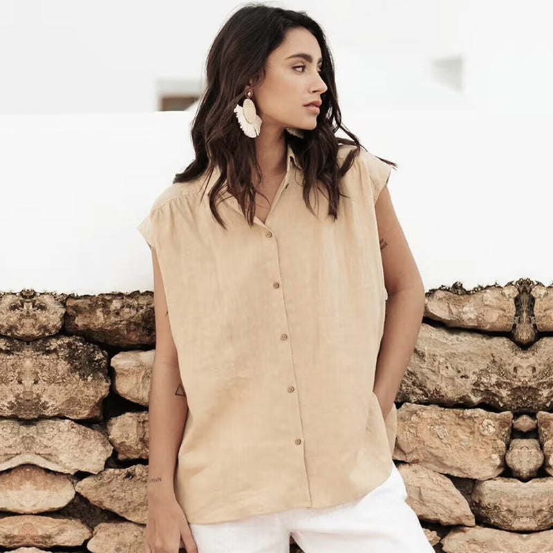 Donna con camicia beige senza maniche, orecchini bianchi, posa davanti a un muro di pietra. Moda estiva, abbigliamento casual elegante.
