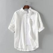 Camicia bianca da uomo in cotone, maniche lunghe arrotolate, colletto alla coreana, design casual elegante, appesa su gruccia.