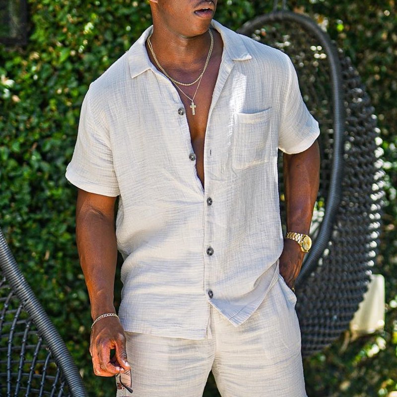 Uomo con camicia di lino beige, collana con croce, orologio dorato, sfondo di foglie verdi. Moda estiva maschile, abbigliamento casual elegante.