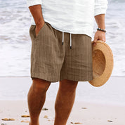 Uomo in spiaggia con pantaloncini marroni a righe, camicia bianca e cappello di paglia. Moda estiva, abbigliamento casual mare.