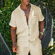 Uomo in camicia di lino beige con pantaloni abbinati, collana e orologio dorato, sfondo verde. Moda estiva casual, abbigliamento maschile elegante.