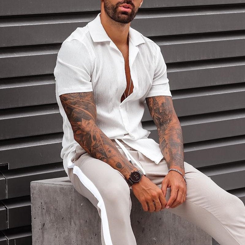 Uomo con camicia bianca a maniche corte, pantaloni beige, orologio elegante e tatuaggi sulle braccia, seduto su panca di cemento. Moda maschile casual.