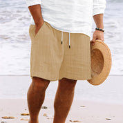 Uomo in spiaggia con pantaloncini beige a righe, camicia bianca e cappello di paglia. Moda estiva, abbigliamento casual mare.