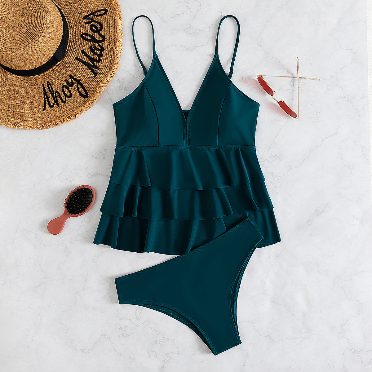 Costume da bagno intero verde con volant su sfondo marmo, accessoriato con cappello di paglia, occhiali da sole rossi e spazzola.