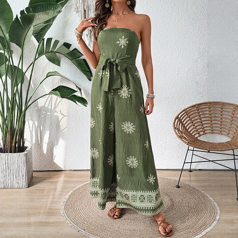 Donna con tuta verde oliva senza spalline, motivo floreale bianco, cintura in vita, sandali marroni, pianta decorativa e sedia in vimini sullo sfondo.
