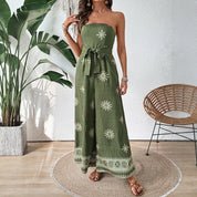 Donna con tuta verde oliva senza spalline, motivo floreale bianco, cintura in vita, sandali marroni, pianta decorativa e sedia in vimini sullo sfondo.