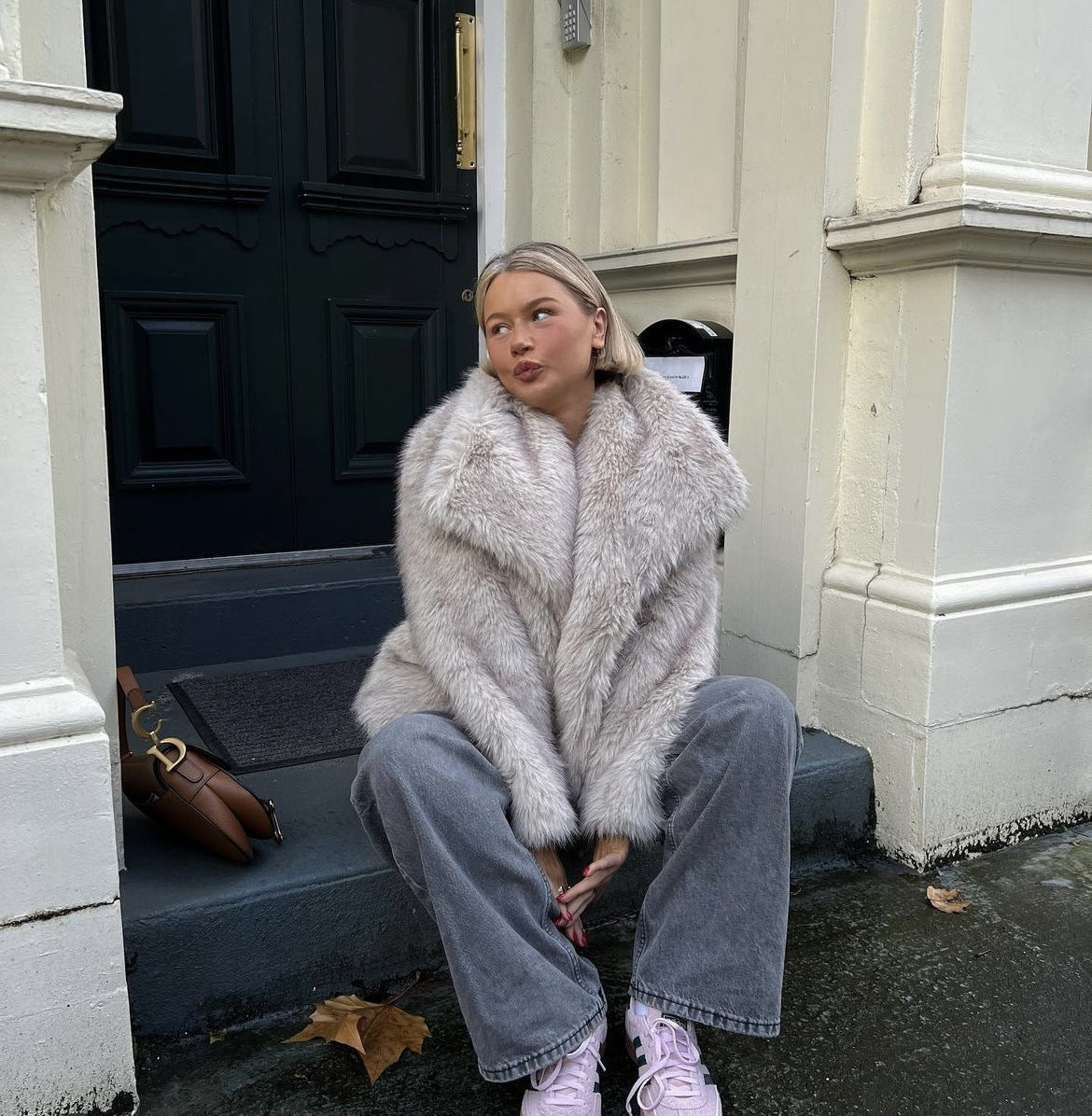 Giovane donna seduta su gradini, indossa cappotto di pelliccia sintetica grigia, jeans larghi e sneakers rosa. Moda invernale urbana, stile casual chic.