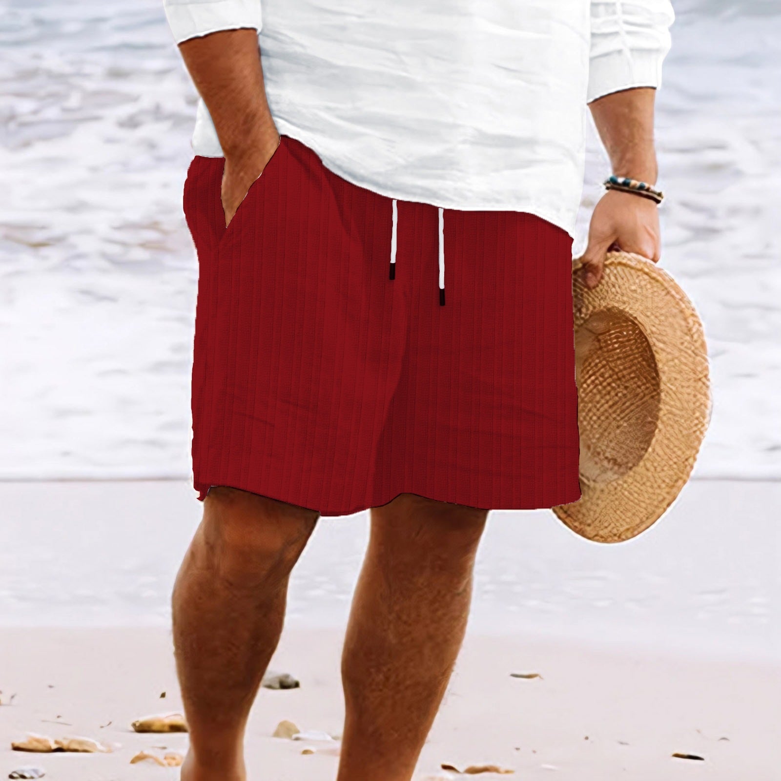Uomo in spiaggia con pantaloncini rossi e camicia bianca, tiene un cappello di paglia. Moda estiva, abbigliamento casual mare.