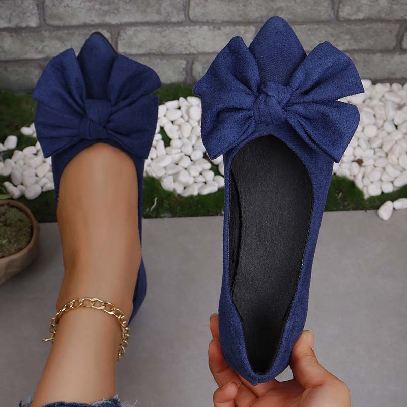 Ballerine blu con fiocco grande, indossate su piede femminile con cavigliera dorata, sfondo di pietre bianche e muro grigio. Scarpe eleganti donna.