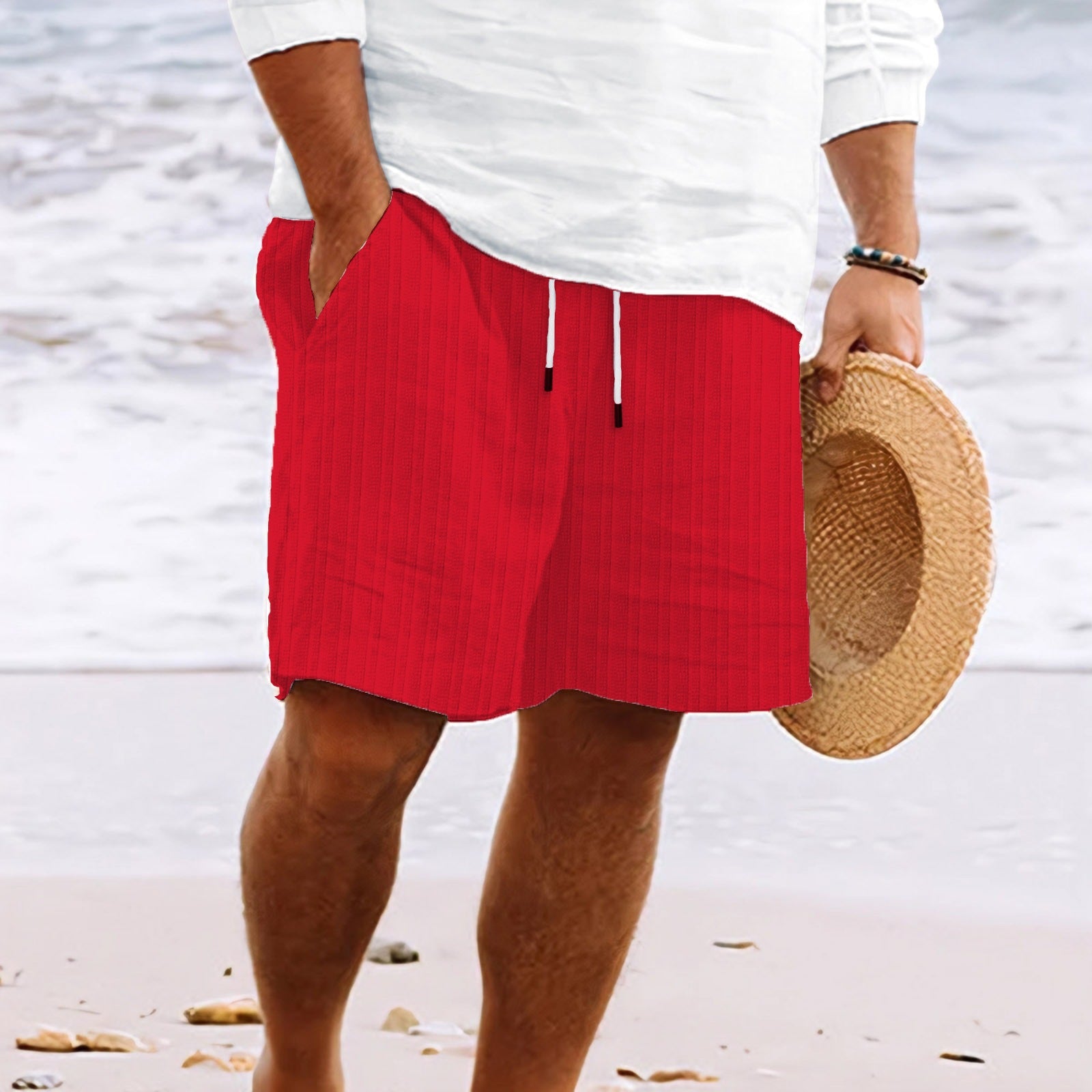 Uomo in spiaggia con pantaloncini rossi casual, camicia bianca e cappello di paglia. Moda estiva, abbigliamento mare, look rilassato.
