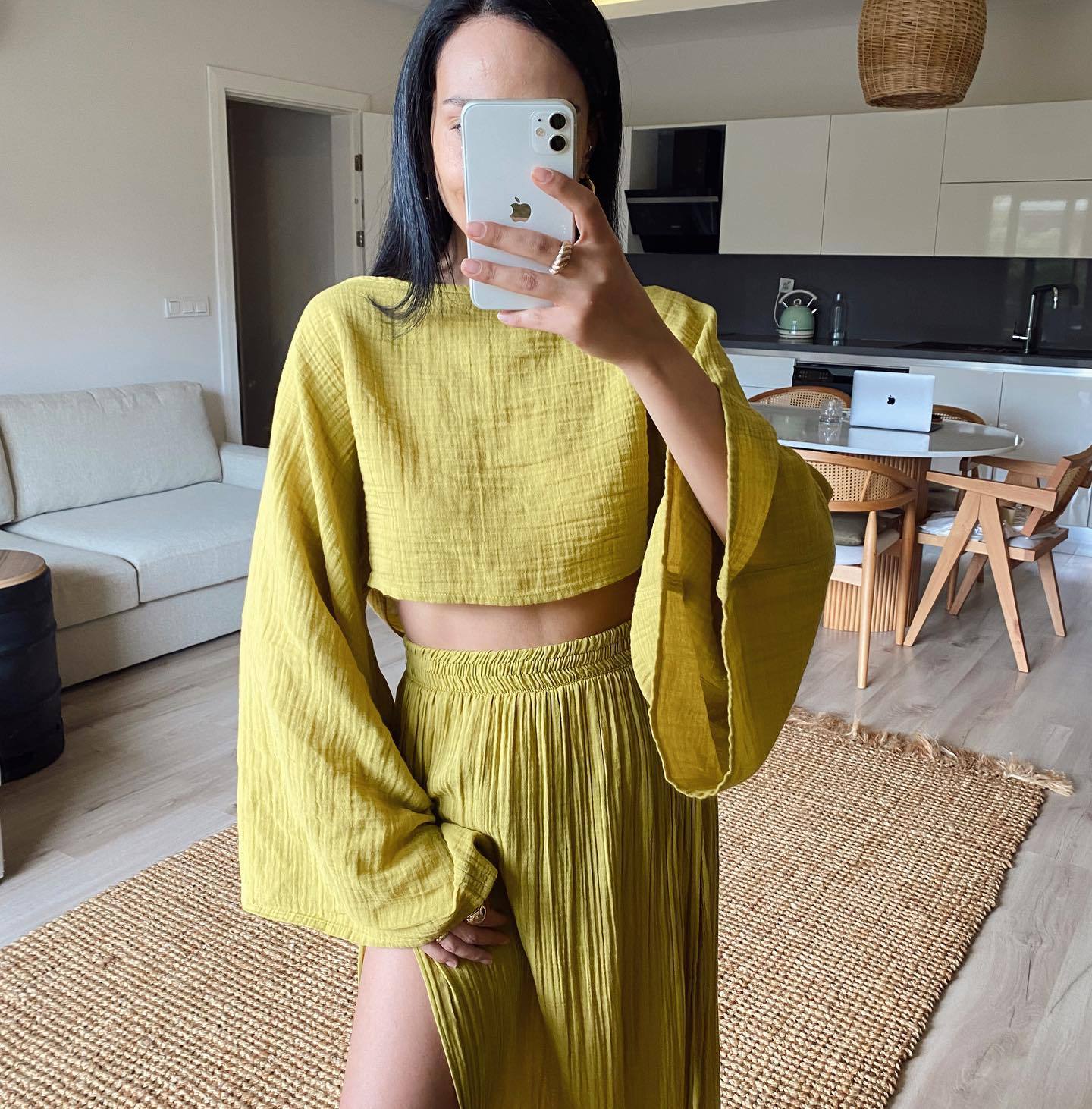 Donna con abito coordinato giallo senape, top a maniche lunghe e gonna plissettata, in soggiorno moderno. Smartphone bianco in mano.