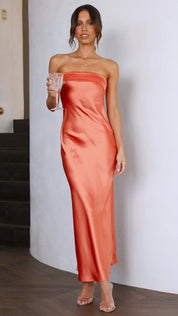 Abito lungo arancione in raso, senza spalline, indossato da una donna con bicchiere di vino. Elegante moda serale, perfetto per eventi formali.