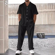 Uomo in completo estivo nero a righe, camicia a maniche corte e pantaloni abbinati, indossa scarpe bianche. Sfondo moderno con parete metallica.
