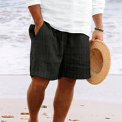 Uomo in pantaloncini neri a righe, camicia bianca, tiene cappello di paglia su spiaggia. Moda estiva, abbigliamento casual mare.