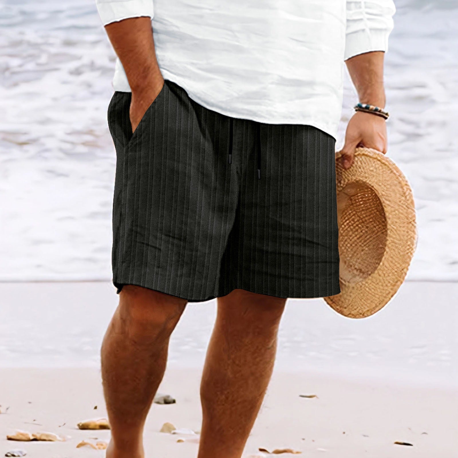 Uomo in pantaloncini neri a righe, camicia bianca, tiene cappello di paglia su spiaggia. Moda estiva, abbigliamento casual mare.
