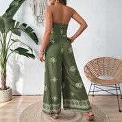 Donna in tuta verde con motivo floreale, senza spalline, in piedi in un interno moderno con piante e sedia in rattan. Moda estiva elegante.