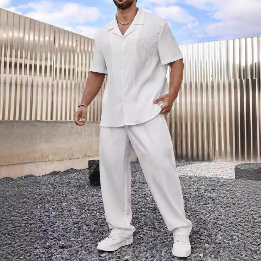 Uomo in completo estivo bianco, camicia e pantaloni larghi, scarpe sportive bianche, sfondo moderno con pannelli metallici e cielo blu.