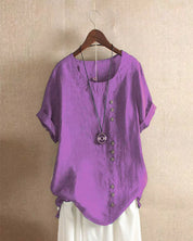 Maglietta viola da donna con bottoni asimmetrici, maniche corte e collana decorativa, appesa su gruccia. Moda estiva casual elegante.