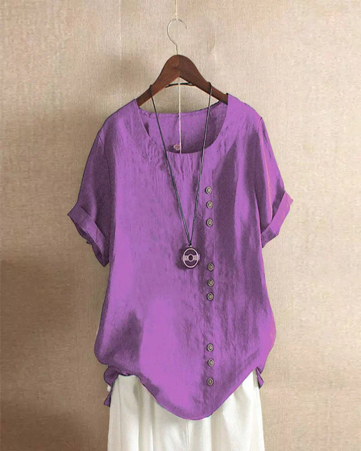 Maglietta viola da donna con bottoni asimmetrici, maniche corte e collana decorativa, appesa su gruccia. Moda estiva casual elegante.