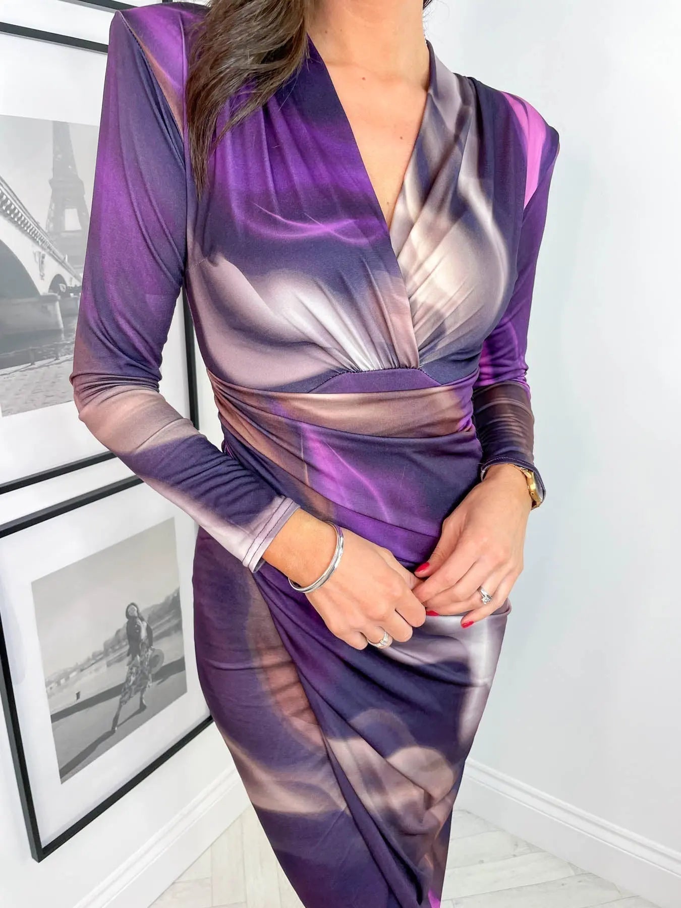 Abito elegante donna viola sfumato, maniche lunghe, scollo a V, indossato da modella. Sfondo con quadri in bianco e nero. Moda femminile chic.
