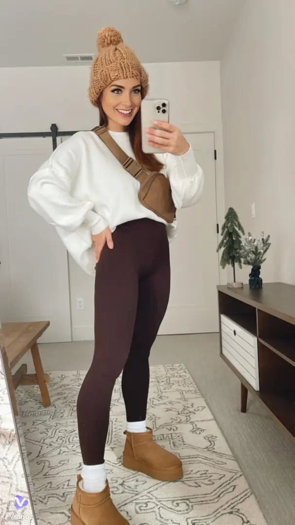 Donna in abbigliamento casual: maglione bianco, leggings marroni, stivali beige, berretto invernale, borsa a tracolla marrone, scatta selfie in camera moderna.