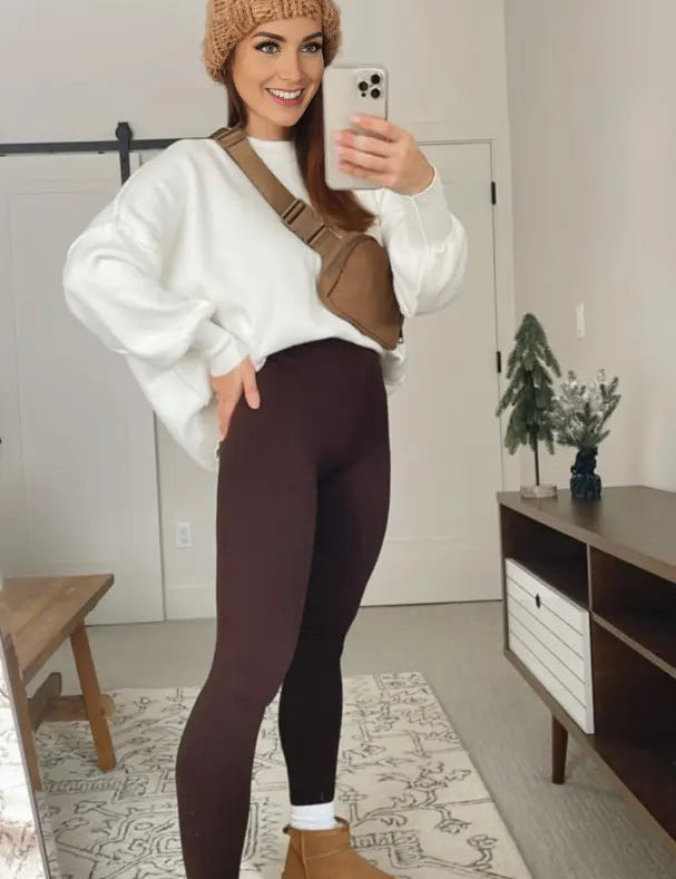 Donna in abbigliamento casual: maglione bianco, leggings marroni, stivali beige, berretto invernale, borsa a tracolla marrone, scatta selfie in camera moderna.