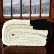 Coperta in pile bianco piegata su tavolo di legno, con finestra che mostra paesaggio invernale innevato. Calda, morbida, ideale per l'inverno.
