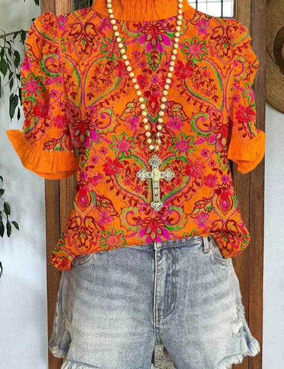 Blusa arancione con motivi floreali vivaci, collana con croce, abbinata a shorts di jeans sfrangiati. Moda estiva, abbigliamento casual donna.