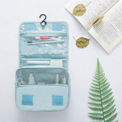 Borsa da viaggio azzurra aperta con gancio, contenente spazzolino Colgate, dentifricio e flaconi. Sfondo con libro aperto e foglie decorative.