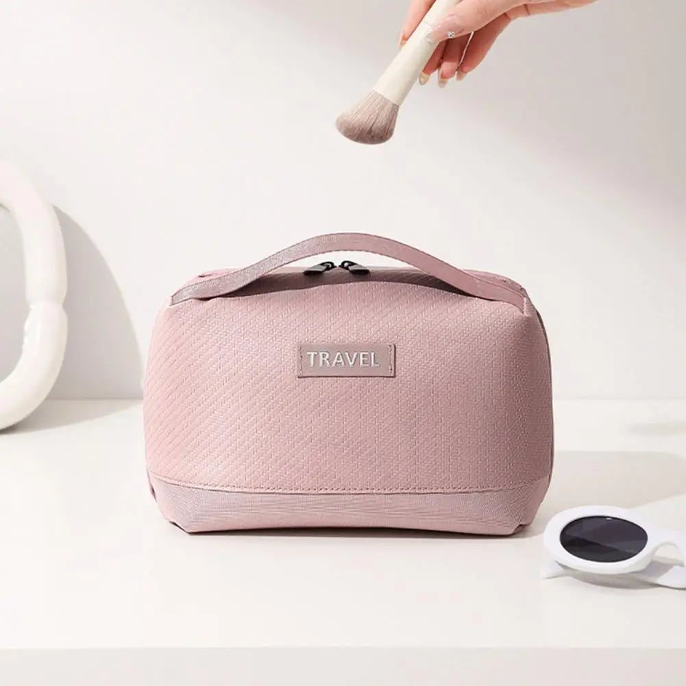 Borsa da trucco rosa con manico, etichetta "TRAVEL", pennello da trucco, occhiali da sole bianchi. Accessorio viaggio elegante e pratico.