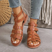 Sandali gladiatore in pelle marrone da donna, con cinturini incrociati e fibbie, indossati con jeans. Scarpe estive eleganti e casual.