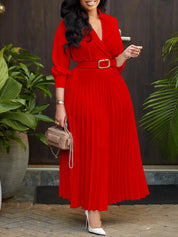Donna in abito rosso plissettato con cintura, borsa a tracolla beige, tacchi bianchi, piante verdi sullo sfondo. Moda elegante, abbigliamento femminile.