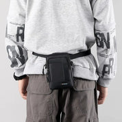 Uomo con marsupio nero in vita, indossa felpa grigia con scritte e pantaloni cargo marroni. Accessorio moda pratico e versatile per uomo.
