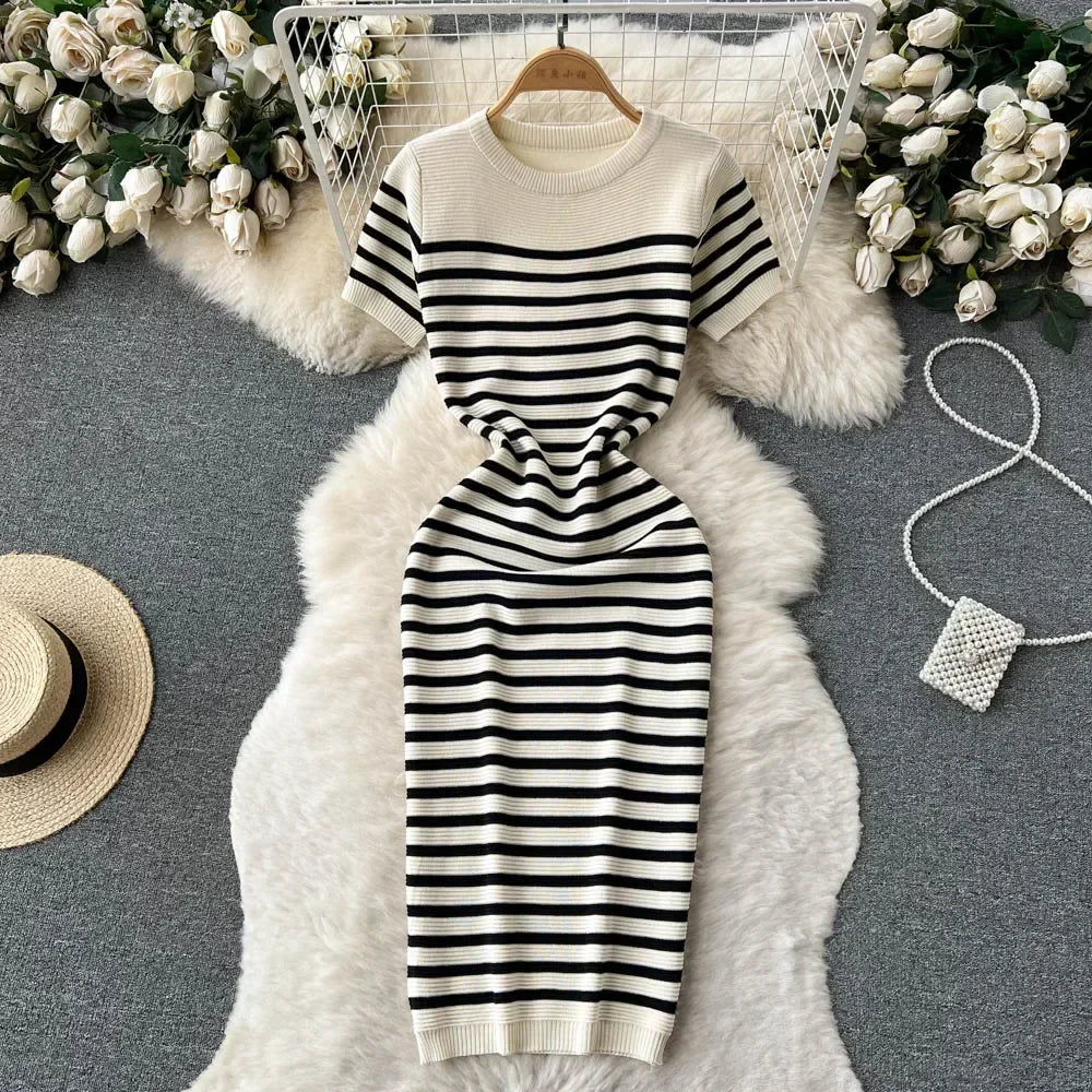 Abito a righe bianche e nere su tappeto bianco, circondato da rose bianche, cappello di paglia e borsa a tracolla. Moda elegante e casual.