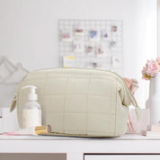 Borsa da toilette trapuntata beige su tavolo bianco, con dispenser sapone e pennello trucco. Accessori bagno eleganti e funzionali.
