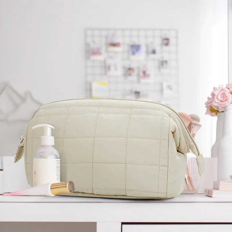 Borsa da toilette trapuntata beige su tavolo bianco, con dispenser sapone e pennello trucco. Accessori bagno eleganti e funzionali.