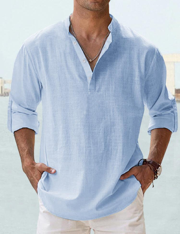 Camicia uomo lino azzurra, maniche lunghe arrotolate, colletto alla coreana, abbinata a pantaloni bianchi. Stile casual estivo elegante.