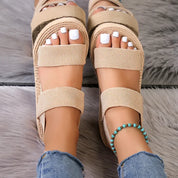 Sandali estivi beige con zeppa, doppio cinturino, indossati su tappeto grigio. Unghie smaltate bianche, cavigliera turchese, jeans strappati.