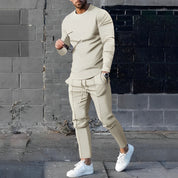 Uomo in tuta sportiva beige, maglia a maniche lunghe e pantaloni abbinati, indossa scarpe bianche, posa casual su sfondo urbano.